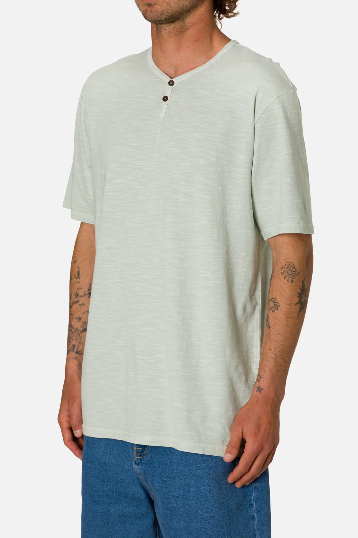 Man wearing a light green henley shirt and blue jeans on a white background