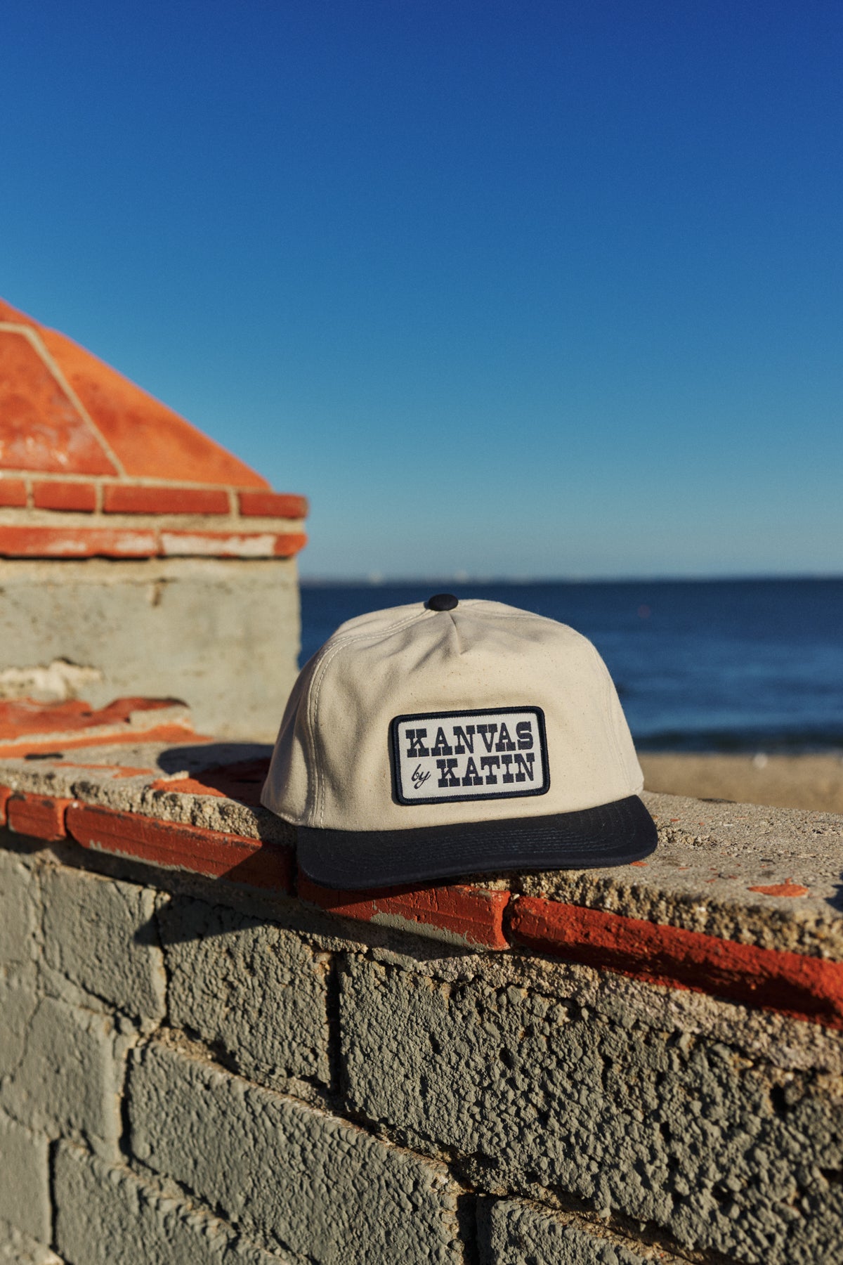 Cap with 'Kanvas &amp; Katin' branding on a brick ledge by the ocean.